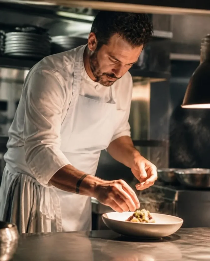 Chef in a white apron plating a dish at the pass of a restaurant kitchen